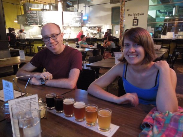 An attractive young man and young woman sitting in a brewpub in front of a sampler of six beers of different colors ranging from dark to light